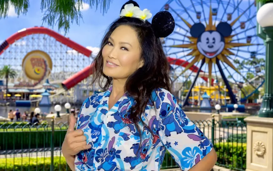 Tia Carrere net worth profile photo at Disney theme park wearing floral dress with Mickey Mouse ears