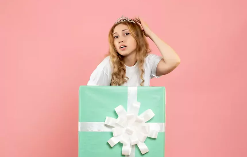 Young woman looking confused at large turquoise mystery box with white ribbon on pink background