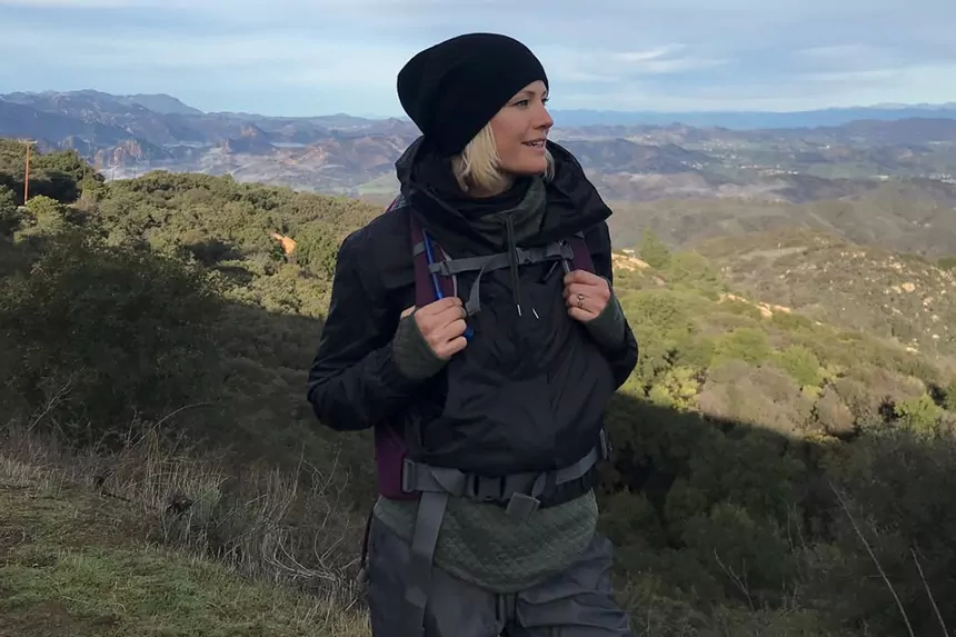 Malin Akerman hiking in the mountains wearing a black jacket and backpack with a scenic valley view behind her